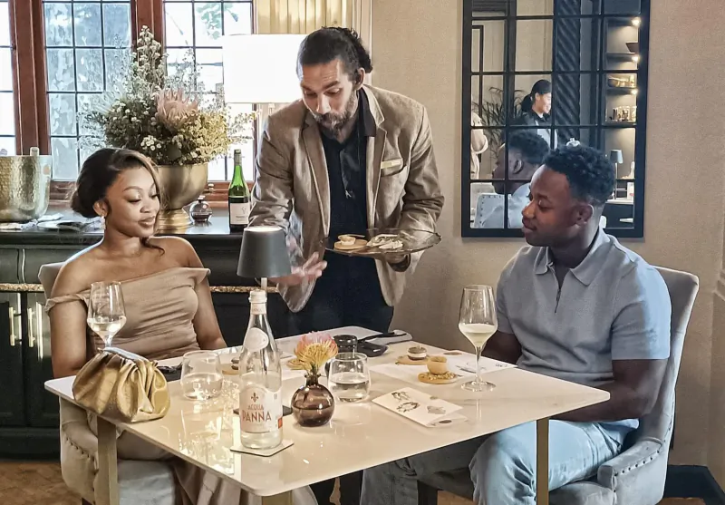 Man in suit serves appetizers to elegantly dressed Black couple at wine tasting table with champagne and flowers.