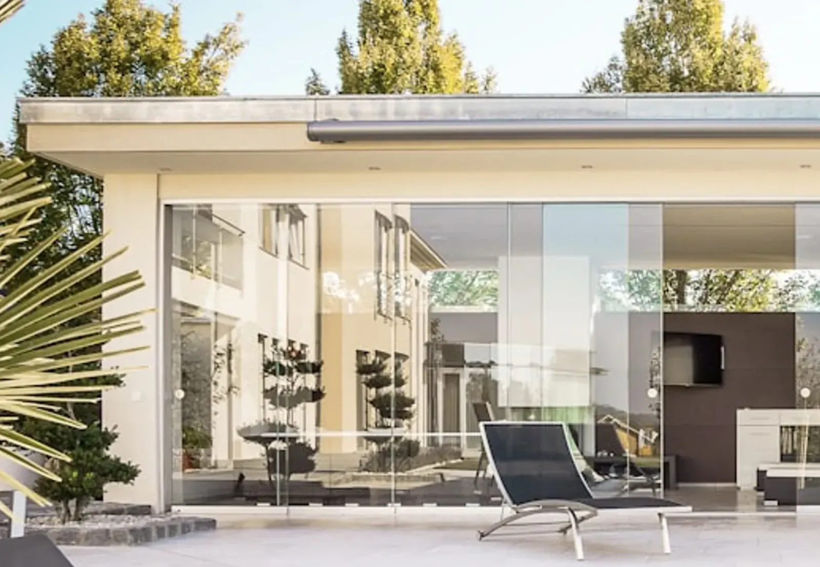Modern luxury home exterior with large glass walls, sleek black lounge chair, fireplace, and potted palms on sunlit terrace