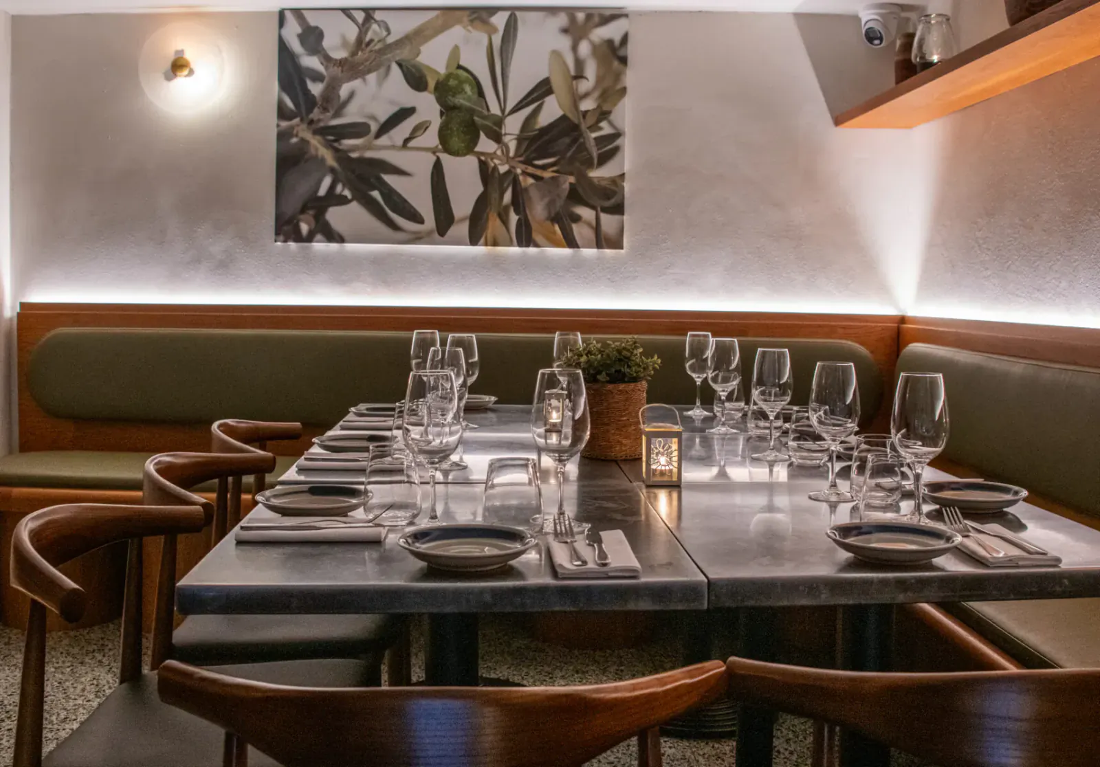 Elegant restaurant interior at Vori, London: long wooden table set with plates, glasses, candles, olive wall art, green banquettes.