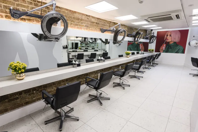 Modern hair salon interior with row of black swivel chairs, hair dryers, mirrors, yellow flowers, and large woman portrait.