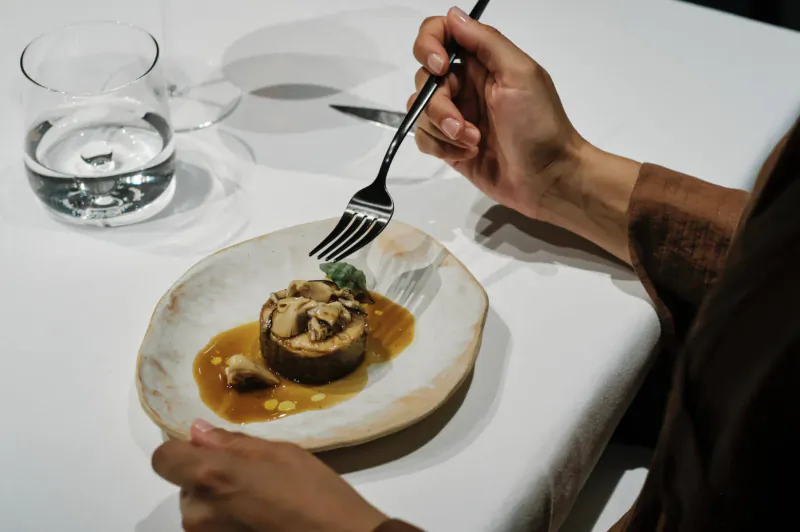 Close-up of hand holding fork over mushroom dish with glossy sauce on white plate at Michelin-starred restaurant table.