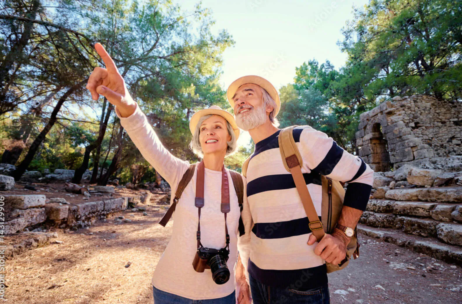 Elderly couple in hats and backpacks point and smile at ancient ruins amid pine trees on sunny path