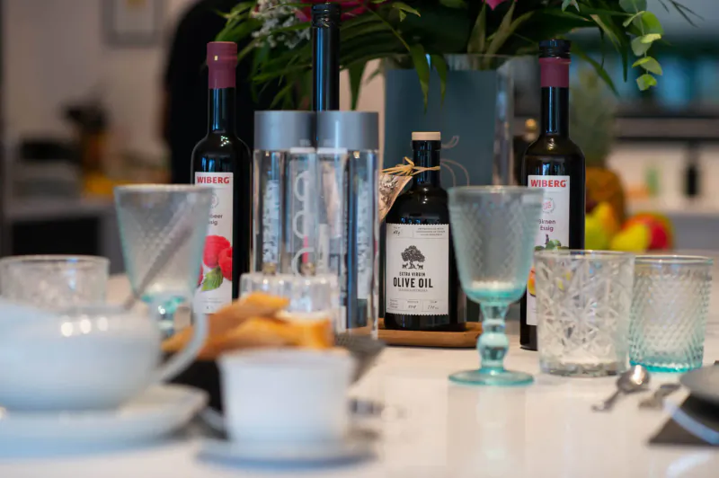 Elegant kitchen table setup with coffee cups, glasses, wine bottles labeled 'Bonaire', small bottle 'Bonaire', flowers, fruit, and pastries.