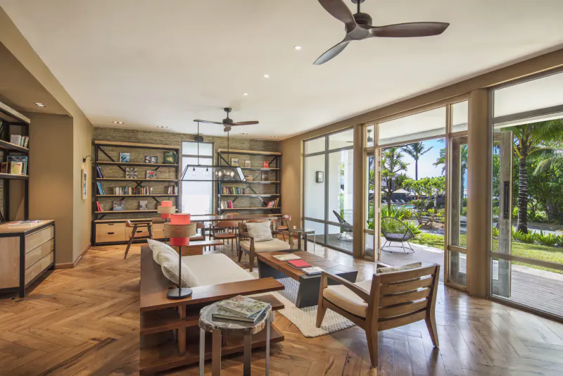 Spacious Anantara Iko Mauritius lounge with wooden bookshelves, low seating, ceiling fans, and tropical garden view through glass doors.