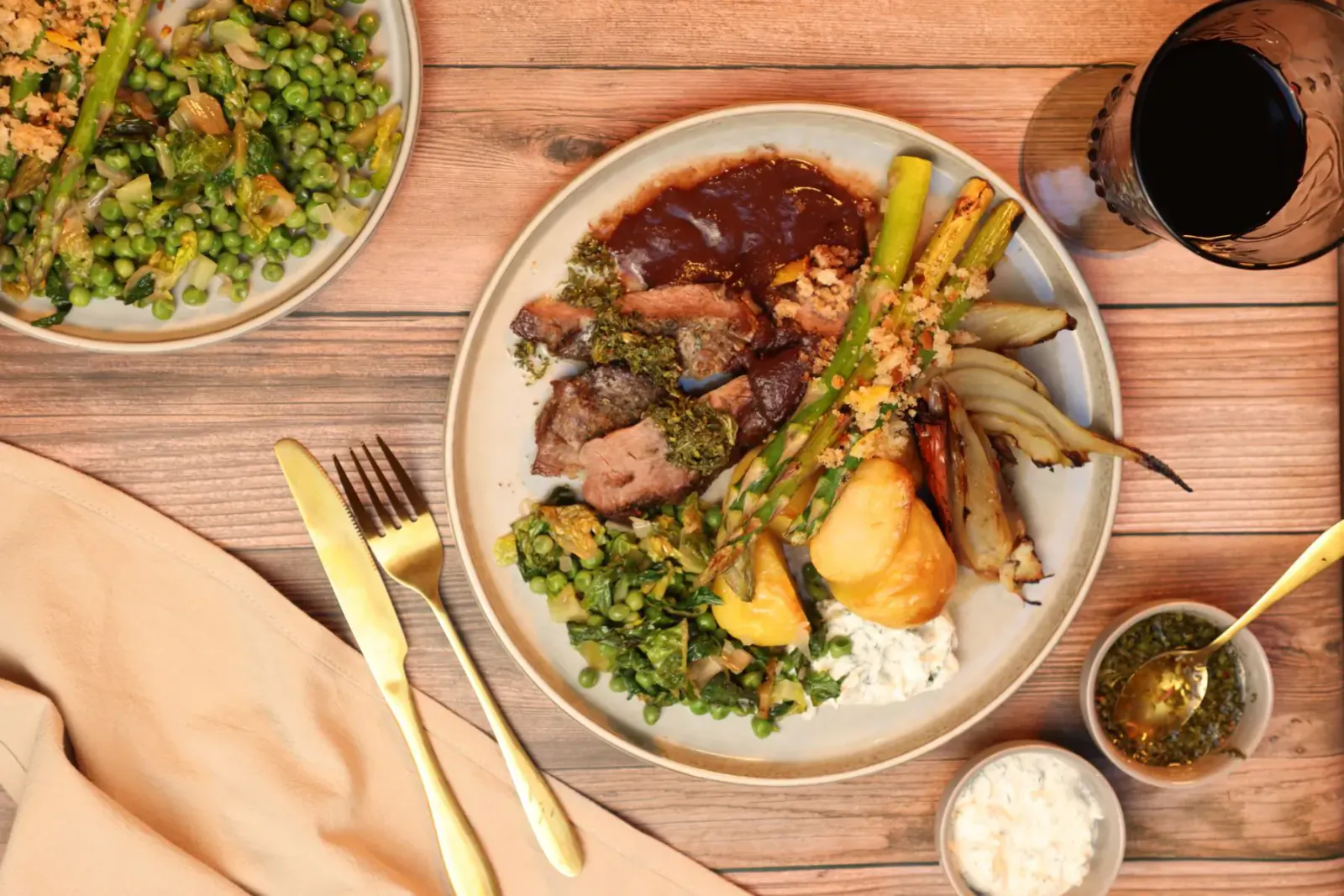 Lamb dish with asparagus, peas, roasted parsnips, and sauce on plate, gold utensils, red wine glass on wooden table