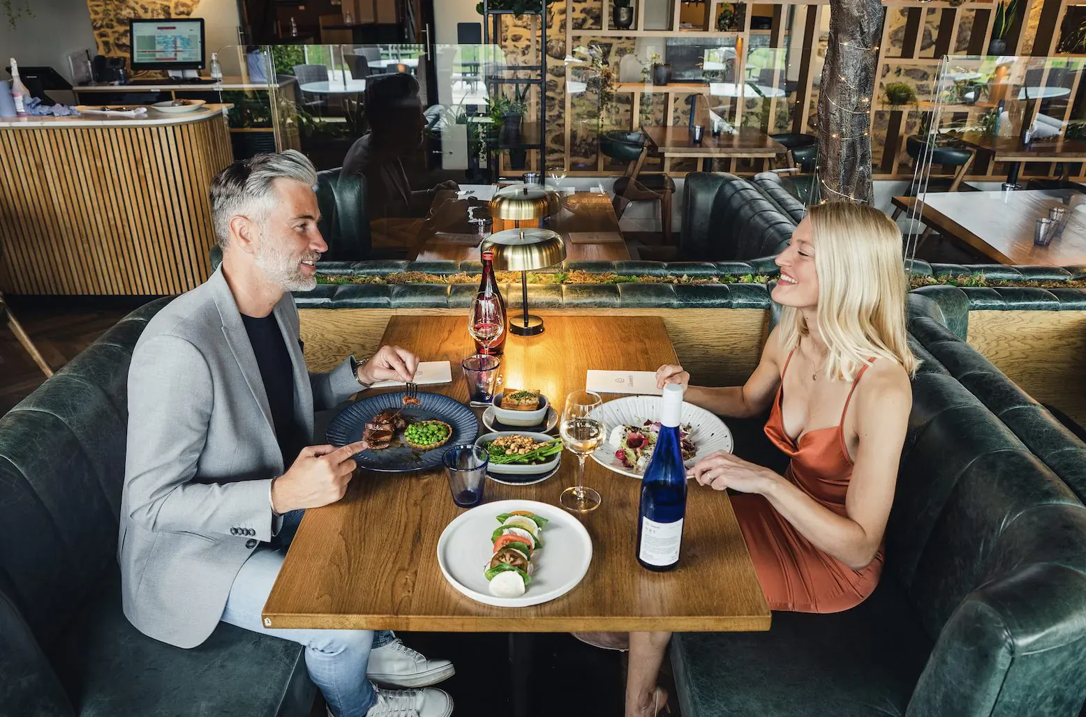 Man and blonde woman smiling, sharing plates of salad and fish at candlelit wooden table in cozy vineyard hotel restaurant.