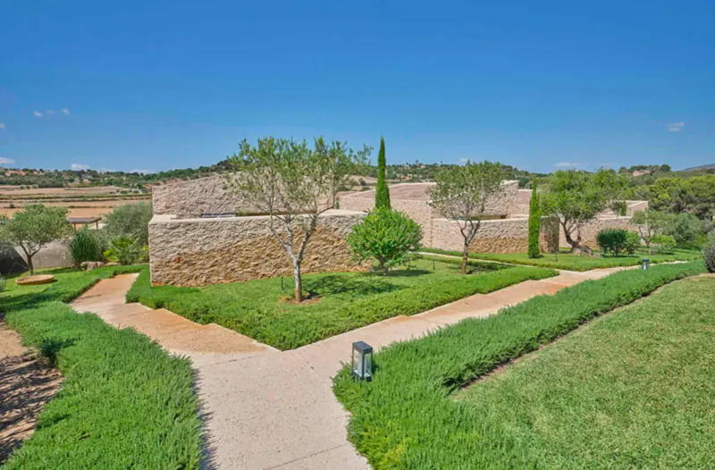 Stone walls of Son Penya Adults Only hotel in Mallorca, with olive and cypress trees, lush gardens, and winding paths under blue sky.