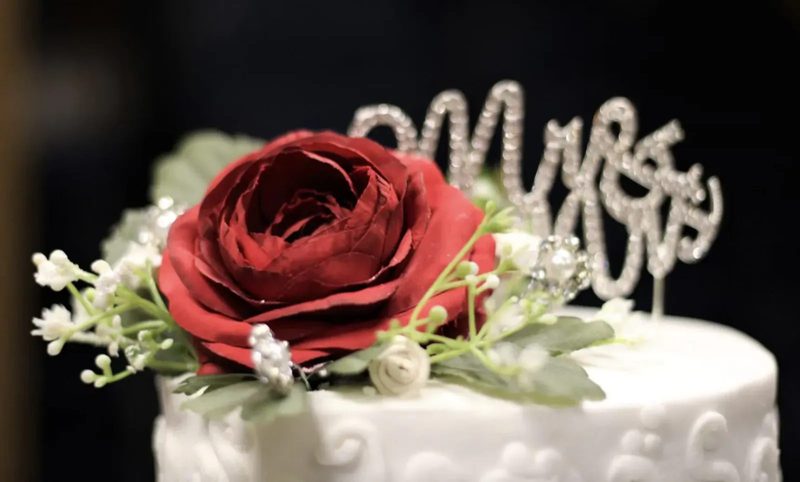 Close-up of white wedding cake topped with red rose, baby's breath, and sparkling 'Mrs' topper.