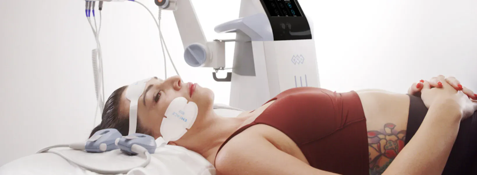 Woman lying on treatment bed receiving EMFACE facial therapy with robotic device, wearing red sports bra and tattoo.