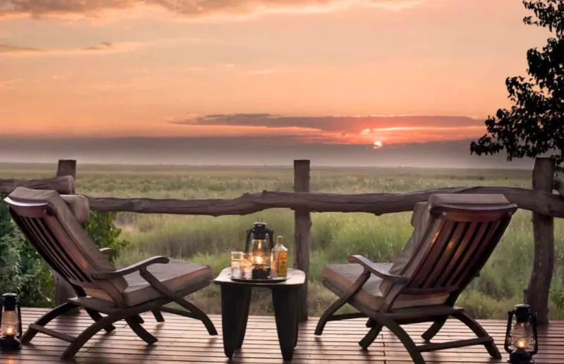 Two wooden chairs and lantern-lit table on deck overlooking African savanna at sunset