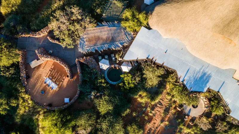 Aerial view of Tswalu's redesigned Tarkuni luxury camp with thatched roofs, deck, pool, chairs amid Kalahari trees.