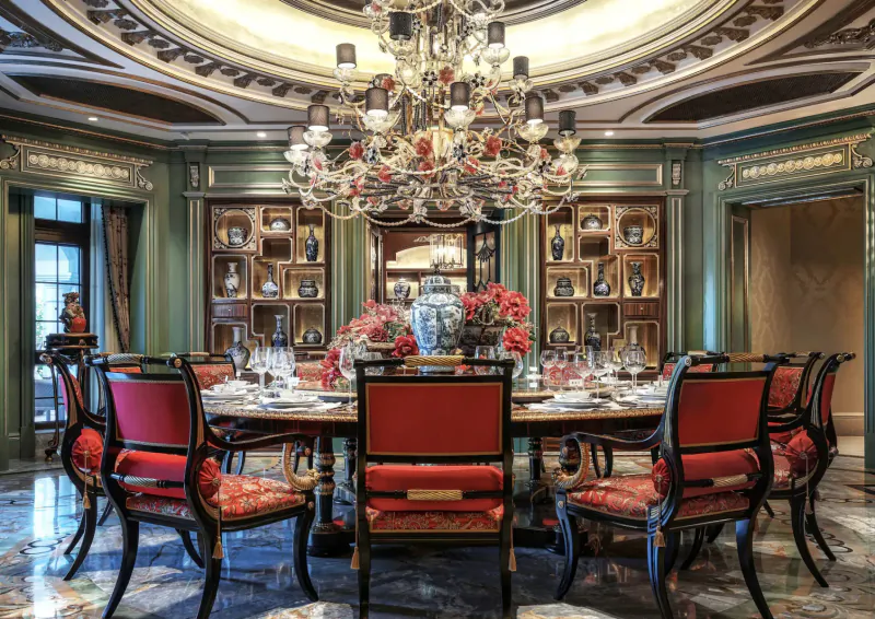 Opulent dining room with grand crystal chandelier, red upholstered chairs around dark wood table, blue-white porcelain vases, and floral centerpiece.