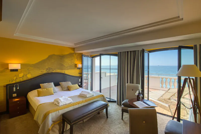 Luxurious hotel bedroom in Saint-Malo with king bed, desk, armchair, and open doors to sea view balcony.