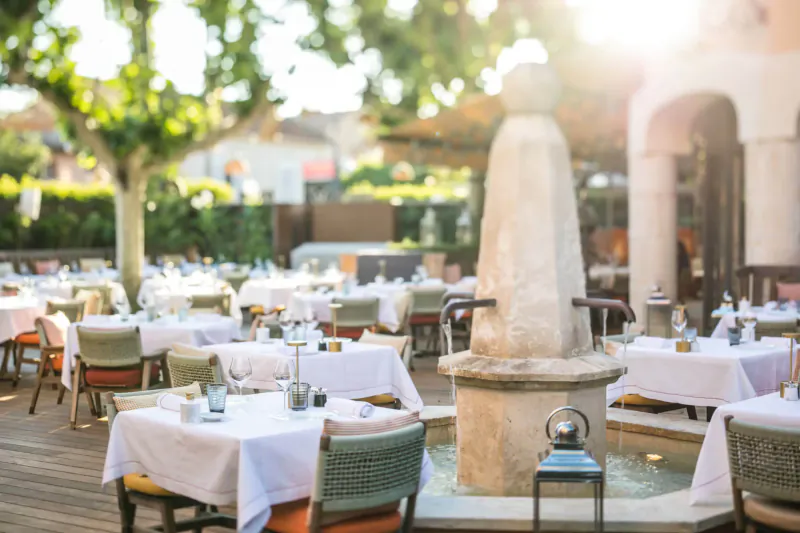 Sunny outdoor terrace at Hôtel Byblos St Tropez with white-clothed tables, stone fountain, trees, and arches.
