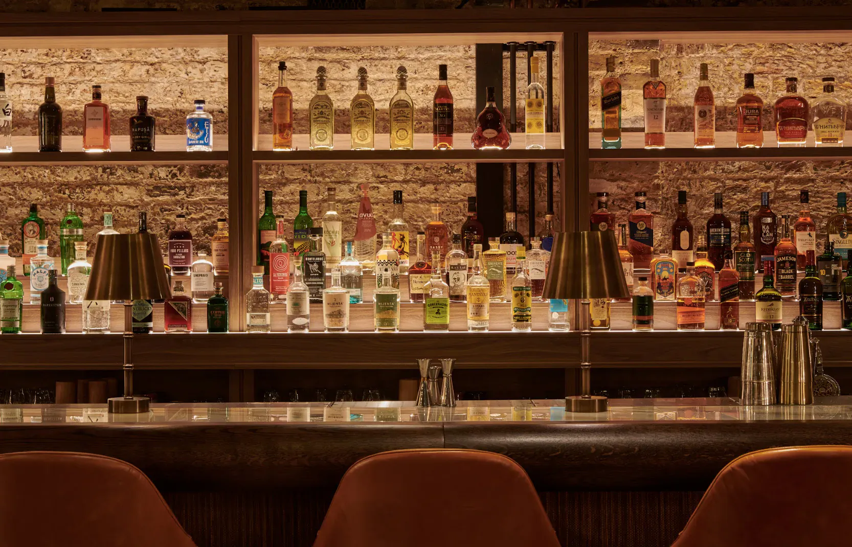 Well-stocked wooden bar backlit with lamps, rows of colorful liquor bottles, bar tools, and empty leather stools at SUCRE bar.