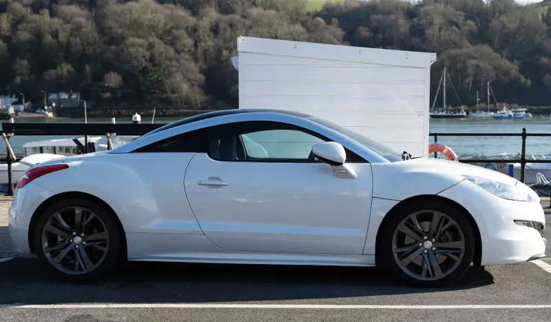 White Peugeot RCZ sports coupe parked by marina with boats, docks, and wooded hills in background.