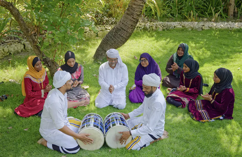 Group in colorful traditional attire playing large drums under palm tree on lush green lawn at Maldives resort