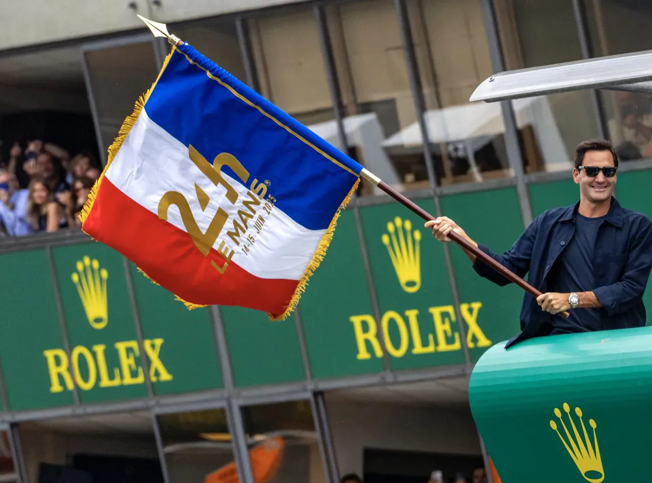 Man in sunglasses and navy jacket holds French 24 Heures du Mans flag on green Rolex podium with crowd.