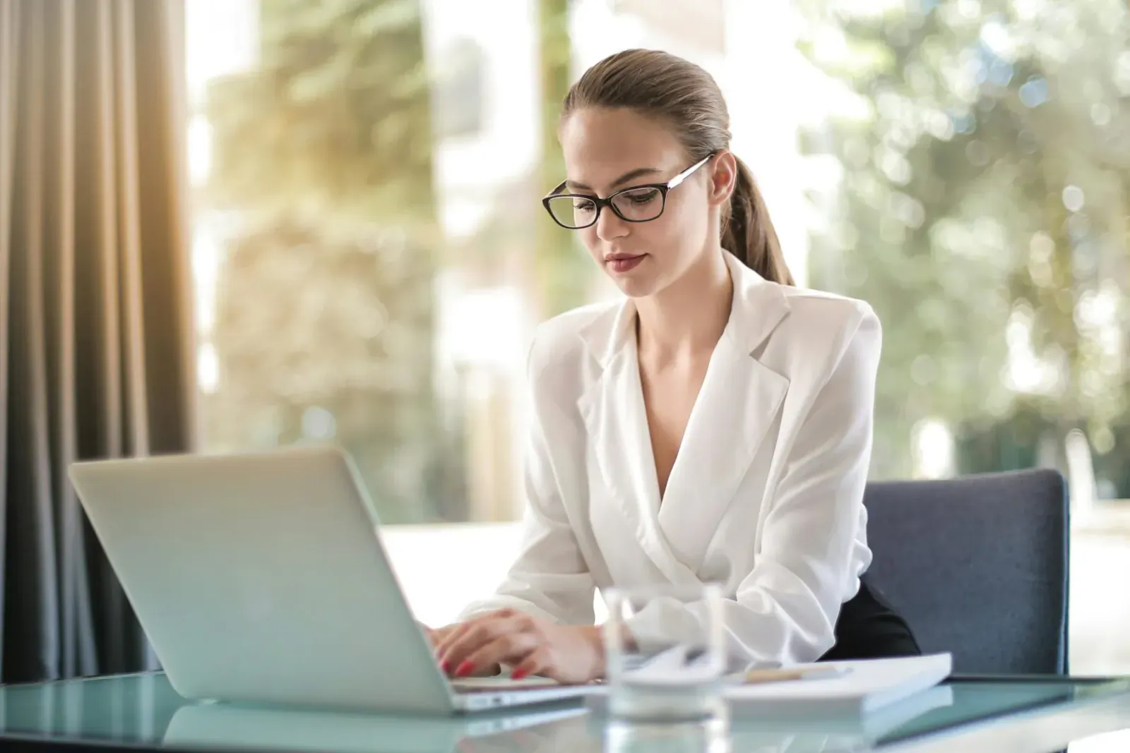 Professional woman with ponytail and glasses typing on laptop at glass desk by sunlit window, wearing white blouse and maxi skirt