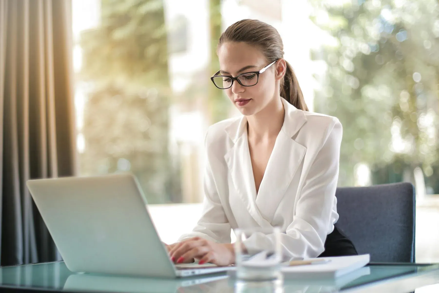 Professional woman with ponytail and glasses typing on laptop at glass desk by sunlit window, wearing white blouse and maxi skirt