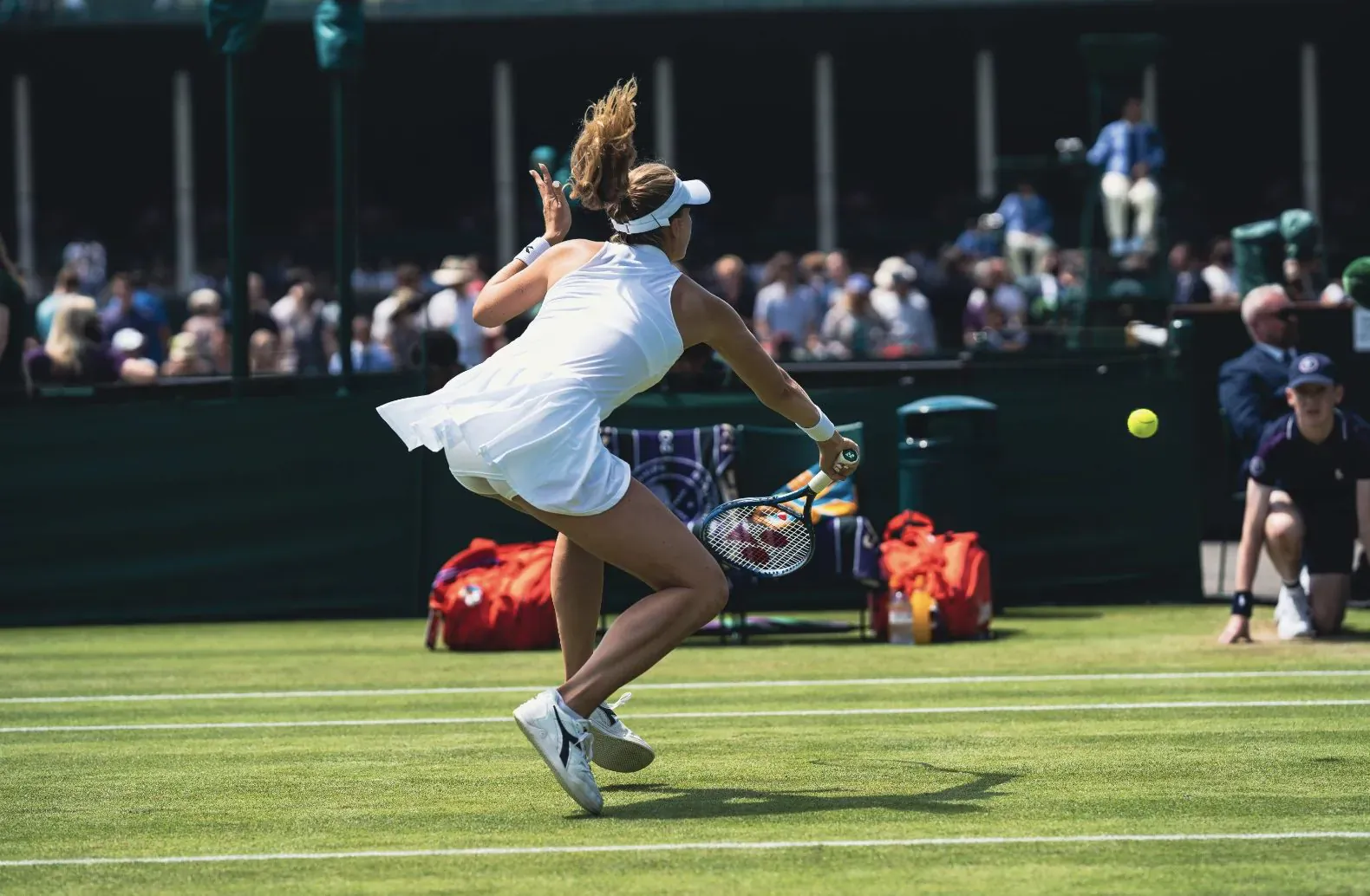 Cornelia Lister in white dress hits yellow tennis ball with racket on grass court at Wimbledon, crowd watches