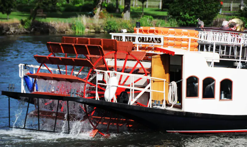 New Orleans paddle steamer boat with red paddlewheels cruising on river near greenery, passengers aboard.