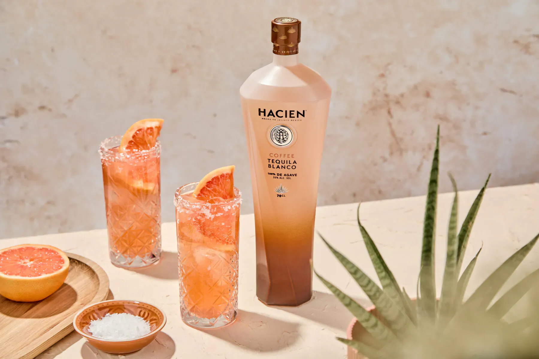 Hacien Tequila bottle with grapefruit paloma cocktails garnished with grapefruit wedges and salt rims on a table with agave plant.