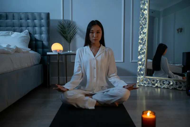 Asian woman in white pajamas sits in lotus pose meditating on yoga mat in elegant bedroom with candle and lamp glow.