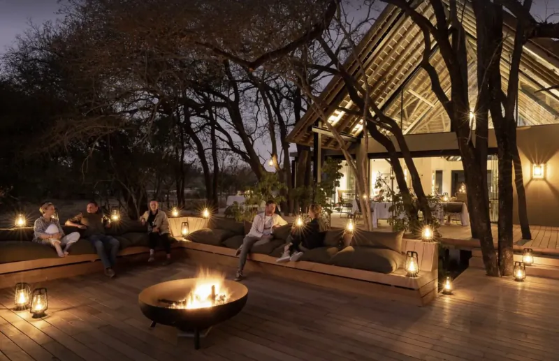 Guests relax around a glowing fire pit on the wooden deck of Monwana Game Lodge at night, lanterns and trees lit.