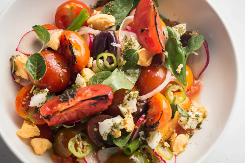 Vibrant Israeli salad in white bowl: grilled tomatoes, feta, olives, radishes, spinach, herbs