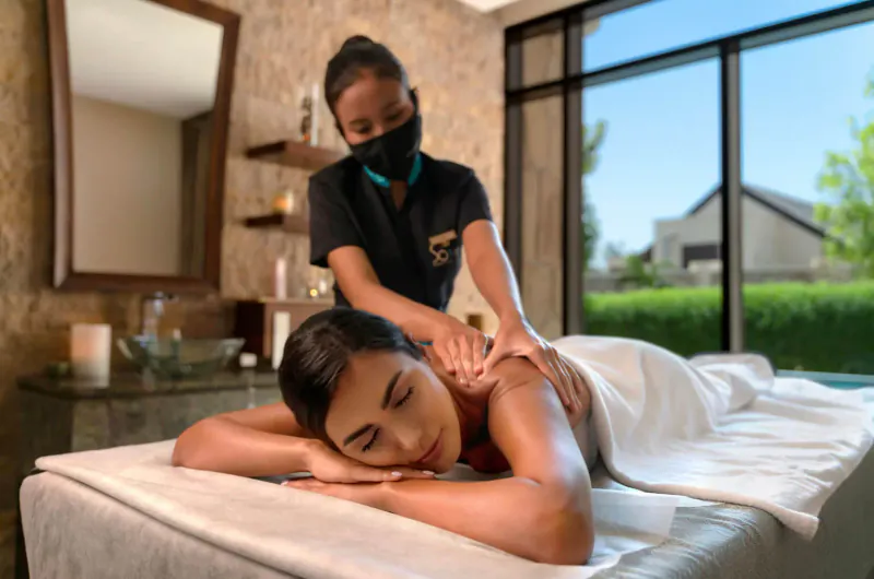 Asian masseuse in black uniform and mask giving shoulder massage to relaxed woman on table in luxurious spa room with garden view.