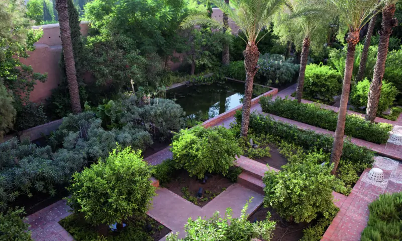 Aerial view of lush Royal Mansour Marrakech medina garden with palm trees, pond, and red brick paths.