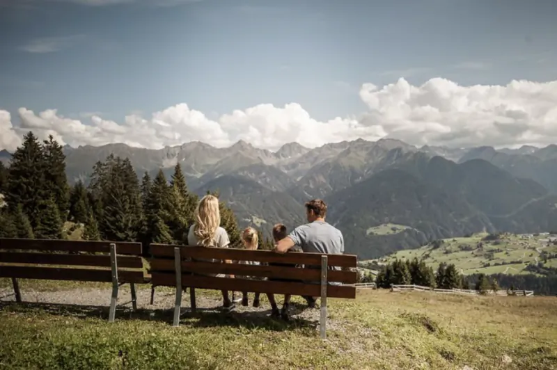 Family with blonde woman, man, and small dog sitting on bench overlooking alpine mountains and valley in spring.