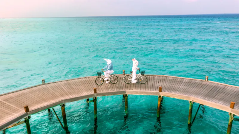 Aerial view of Easter Bunny and Chef Thomas Alphonsine in white outfits biking on curved wooden pier over turquoise sea at Hideaway.