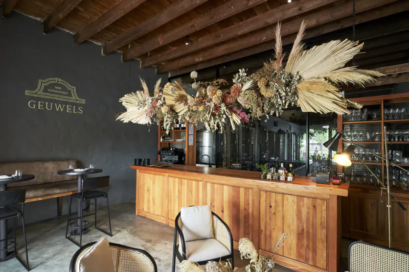 Cozy bar at Geuwels with wooden bar, pampas grass hanging decor, stools, chairs, and 'Geuwels' logo on wall