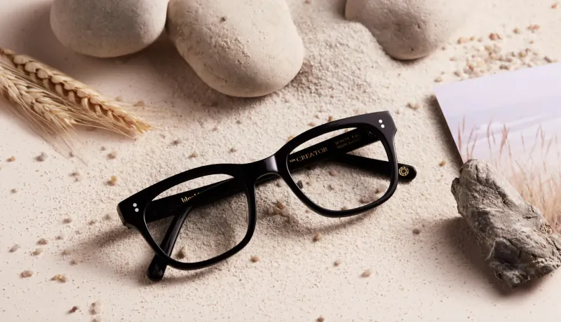 Black rectangular spectacles on sandy surface with pebbles, wheat stalks, and sand, for Bloobloom Pair for Pair scheme.