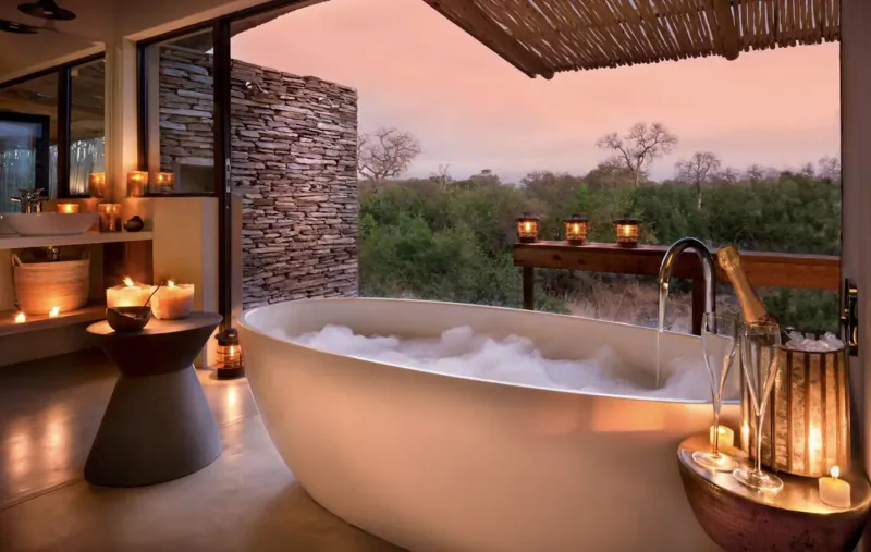 Luxurious outdoor bathtub with bubbles and champagne on ice at sunset, overlooking African savanna bushveld.