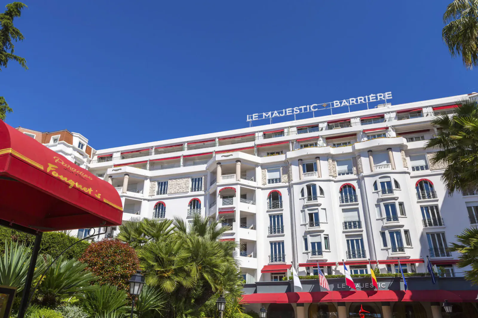 Hôtel Le Majestic Barrière in Cannes, grand white building with red awnings, palm trees, and blue sky.
