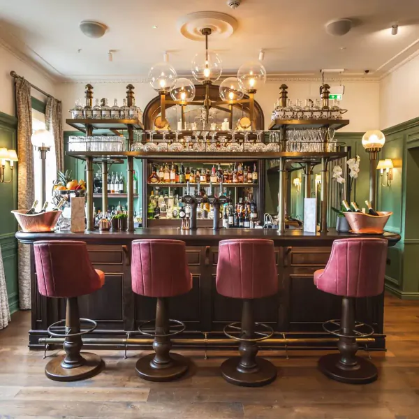Elegant green bar at Hotel Indigo Bath with ornate chandelier, extensive liquor shelves, red stools, and champagne bottles.