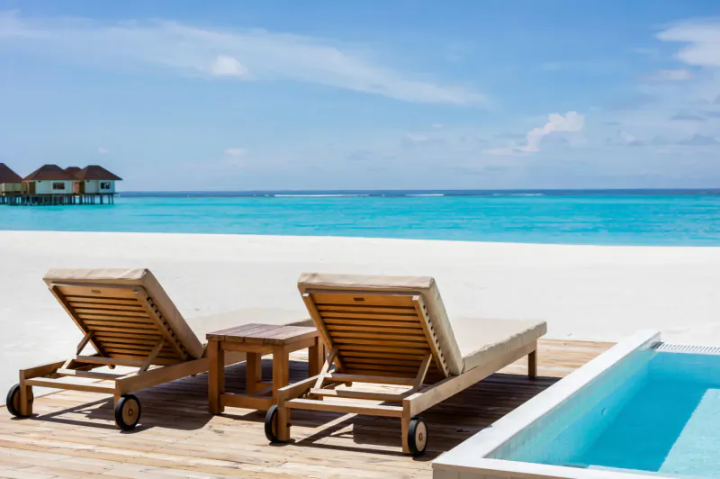 Two wooden sun loungers with cushions on a wooden deck beside an infinity pool, white sand beach, turquoise sea, and overwater villas under blue sky.
