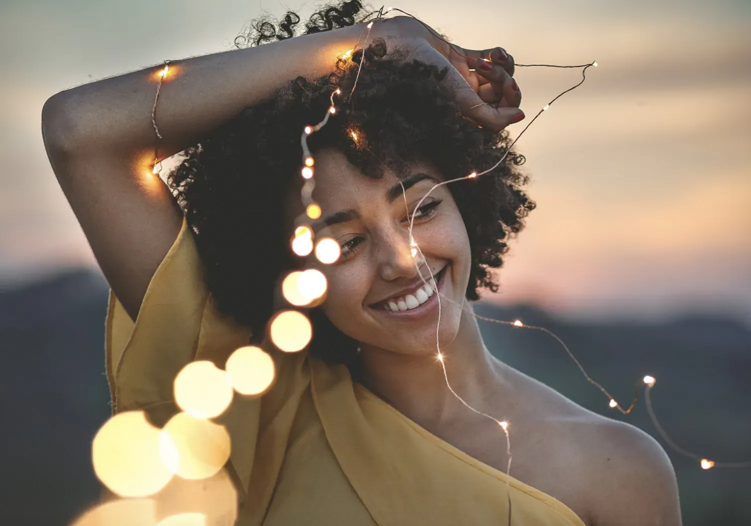 Smiling Black woman with curly hair holding fairy lights, wearing yellow dress at sunset mountains