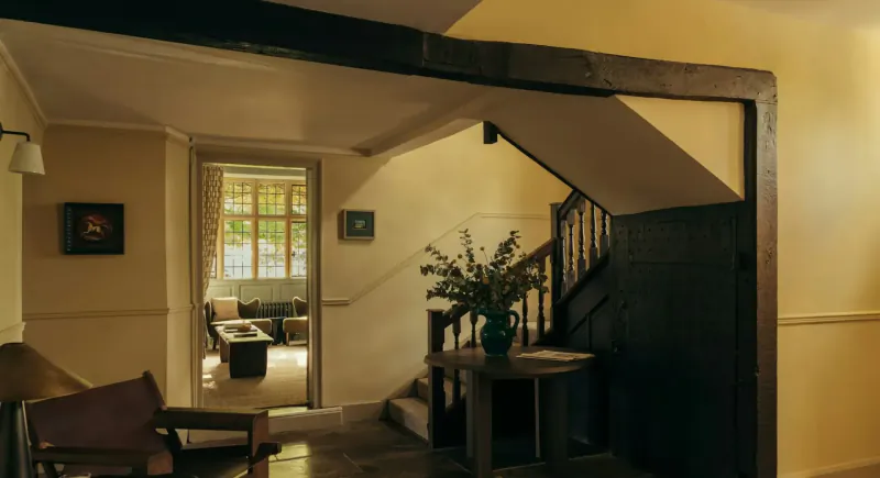 Warmly lit cozy interior at Hyll with wooden staircase, green vase of flowers on table, armchair, and sunlit room beyond.