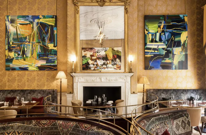 Elegant Home House gallery lounge with ornate gold-framed paintings, colorful abstract art, fireplace, curved seating, and lamps.