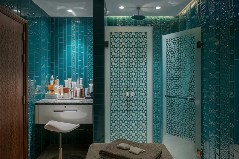 Luxurious turquoise-tiled spa bathroom with skincare products on vanity, white stool, folded towels, and latticed doors.