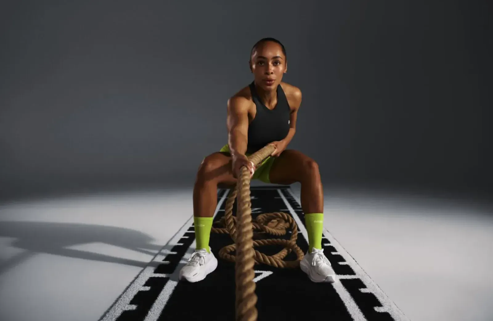 Fit Black woman in black tank top and neon green socks squats pulling thick rope on gym track