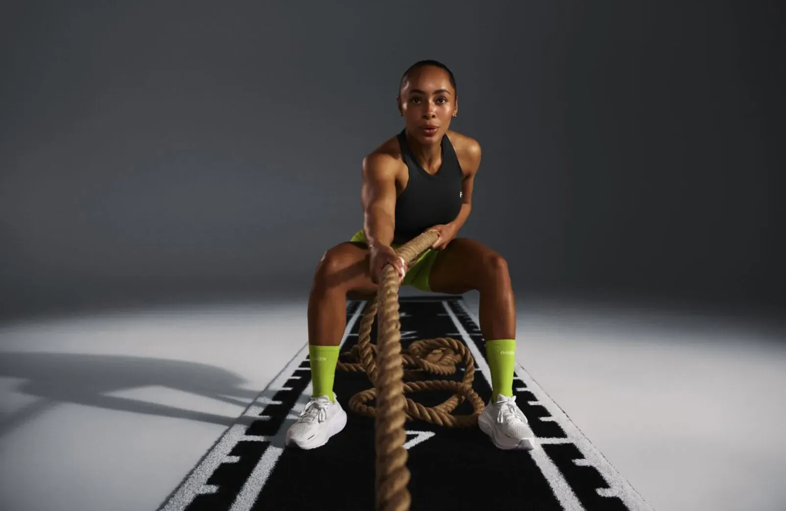 Fit Black woman in black tank top and neon green socks squats pulling thick rope on gym track