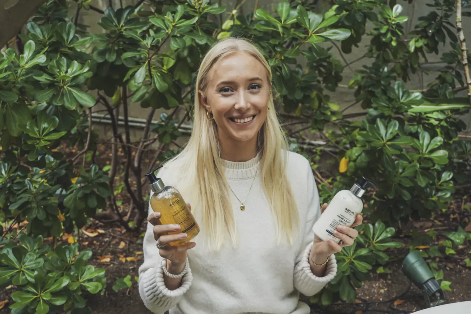 Sine Brynestad Stokke, smiling blonde woman in white sweater, holds amber and white Elm Organics bottles amid lush green plants.