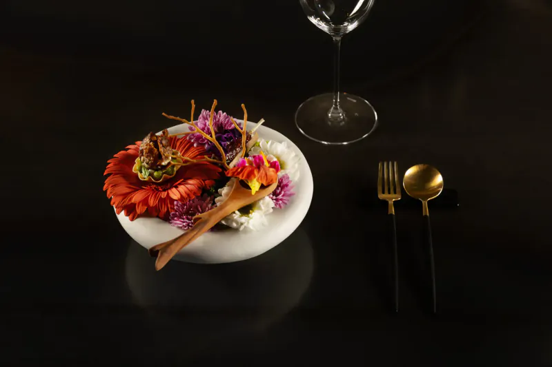 White bowl of vibrant edible flowers and garnishes with chopsticks, beside gold fork and knife, wine glass on black table