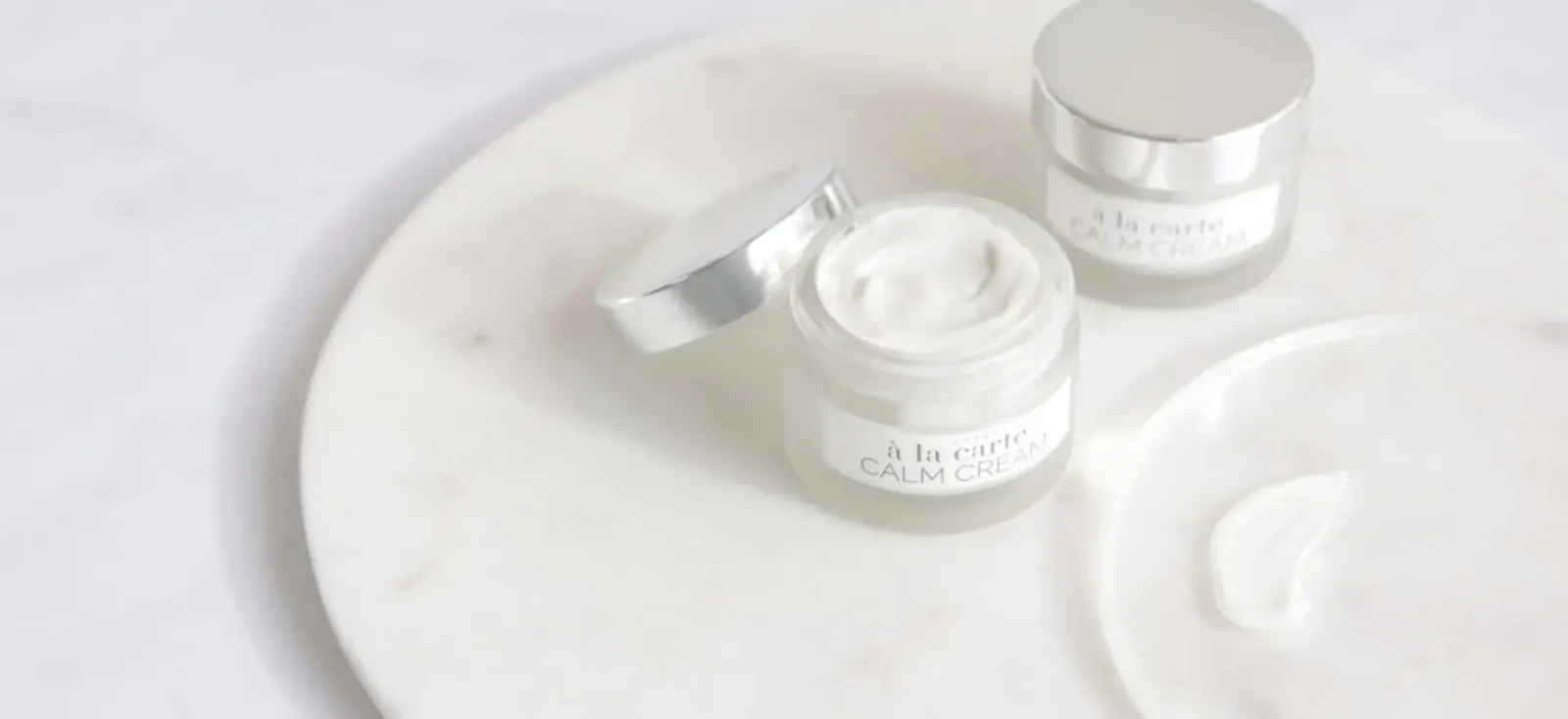 Two open Calm Cream jars with white cream on marble surface, one with cream scoop