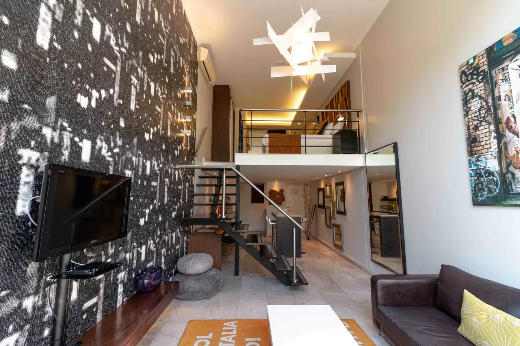 Modern loft room at Hippo Boutique Hotel with black patterned wall, metal staircase to mezzanine, leather sofa, TV, and orange rug.